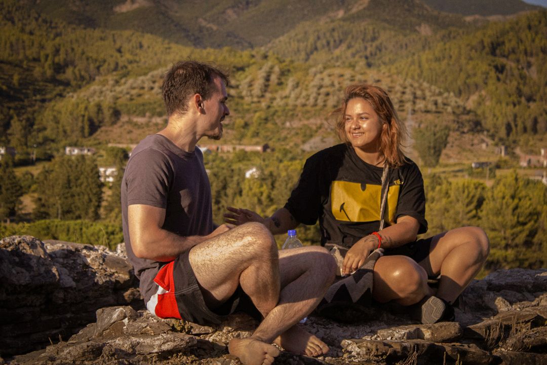 A traveler and a local sit talking on a hilltop at sunset.