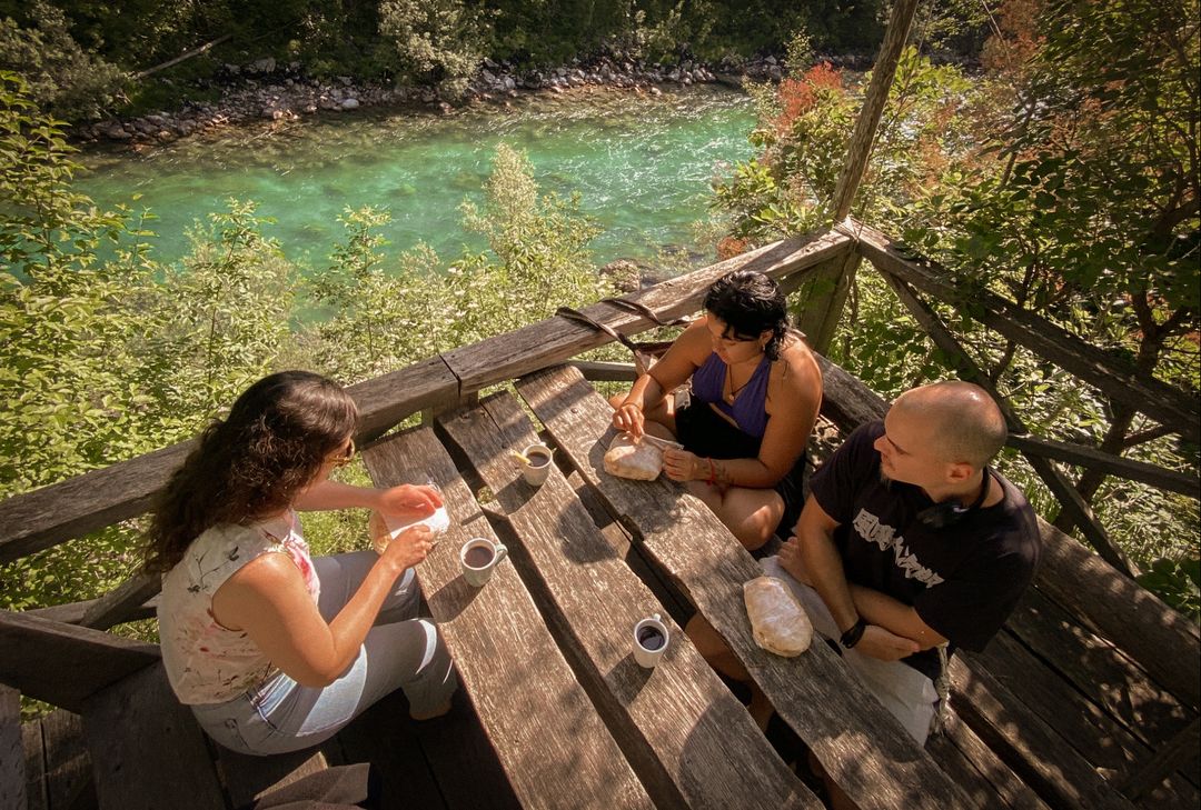 Three friends share bread and coffee at a wooden table overlooking a river.