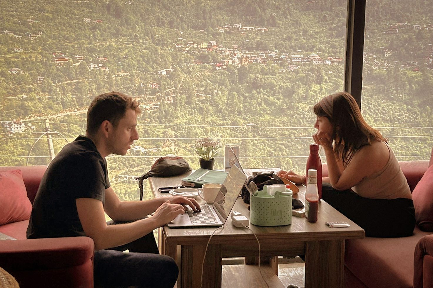 A traveler and a local talk over a laptop at a table overlooking a hillside town.