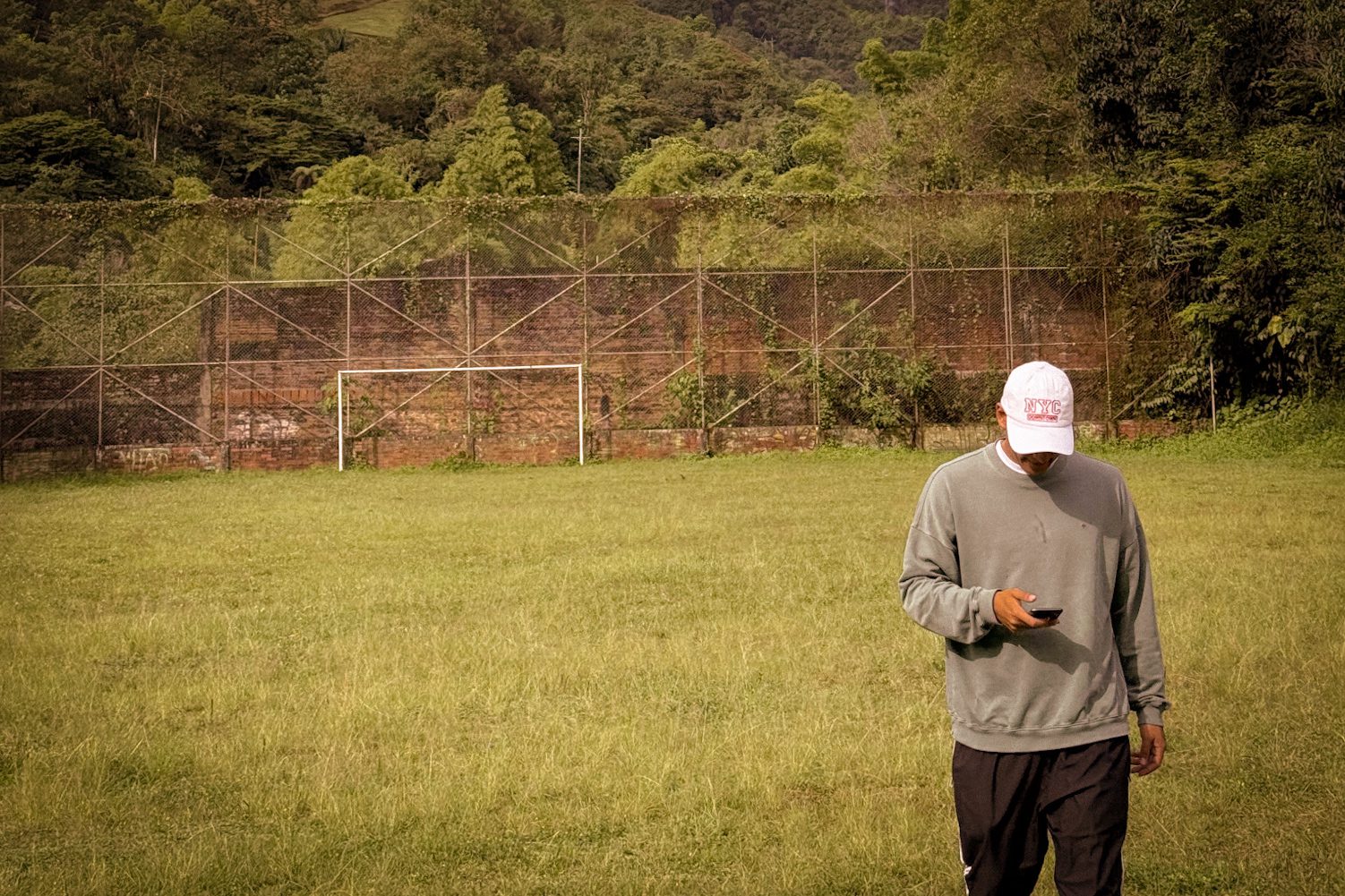 A traveler checks his phone while walking across an open field with a soccer goal in the background.
