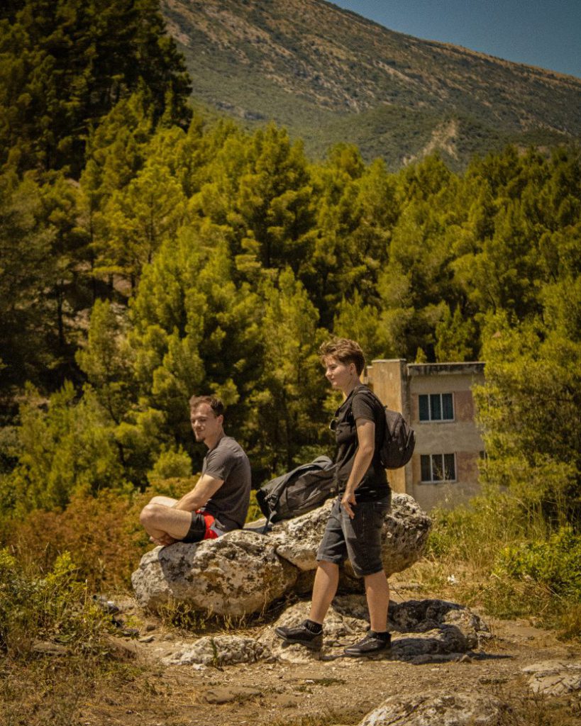 Two travelers pause on a rocky outcrop, with pine trees and mountains rising behind them.