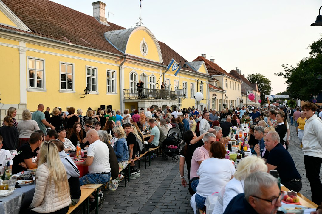 Dozens of people sharing a meal together along one long wooden table set up in a historic town square